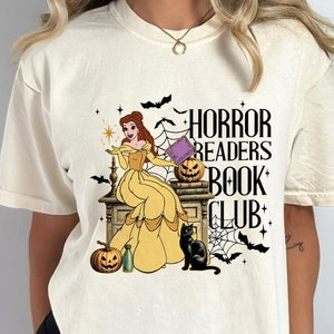 May include: Cream-colored t-shirt with a graphic featuring a woman in a yellow dress, pumpkins, bats, and a black cat. The text "HORROR READERS BOOK CLUB" is displayed in black. A Halloween-themed design.