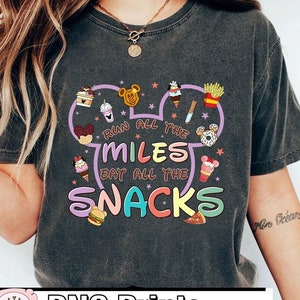 May include: A gray t-shirt with a purple Mickey Mouse head design. The design includes various food and drink items, and the text "Run all the miles eat all the snacks".