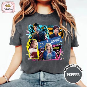 May include: Dark gray t-shirt featuring a colorful graphic of characters from "Zombies" with the names Zed, Victor, Addison, and Nova. The shirt has the words "Zombies" and "Comfort Colors Pepper" printed on it.