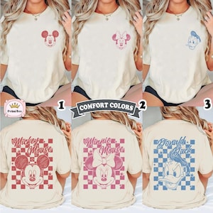 May include: Cream-colored t-shirts featuring Mickey Mouse, Minnie Mouse, and Donald Duck designs. Each shirt has a character graphic on the front and back. The text "COMFORT COLORS" is displayed on a banner.