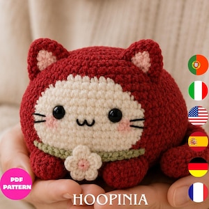 Peut inclure: Une peluche chat crochetée rouge foncé, ronde, avec un visage crème, des yeux en plastique noir et une bouche brodée. Elle présente des joues roses, un collier vert clair avec une petite fleur crème et des oreilles roses. Texte PDF PATTERN HOOPINIA.
