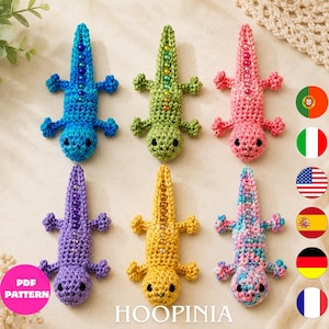 May include: Six crocheted lizard toys arranged in two rows. The lizards are blue, green, pink, purple, yellow, and a multi-colored mix. Each lizard features a row of small beads along its back and two black eyes. A pink circle with the text PDF PATTERN is visible.