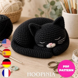Könnte beinhalten: Eine schwarze gehäkelte Katzenfigur in zusammengerollter Schlafposition mit weiß gestickten Schnurrhaaren und einer kleinen rosa Nase. Die Katze ist mit einem dichten Maschenmuster gefertigt und hat spitze Ohren auf einer Oberfläche.