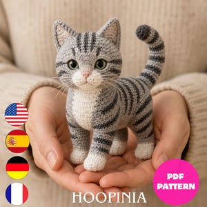 Puede incluir: Una figura de gato atigrado tejida a ganchillo en gris y blanco con rayas gris oscuro, ojos verdes y nariz rosa. El gato tiene la cola levantada y patas blancas. El texto "HOOPINIA" y "PDF PATTERN" aparecen junto a cuatro iconos de banderas.