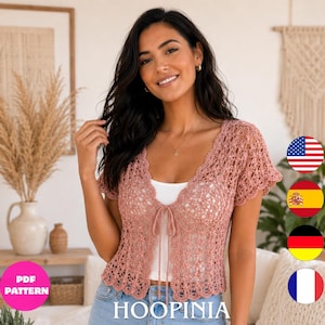 Può includere: Un cardigan all'uncinetto color rosa polvere con maniche corte, bordo smerlato e chiusura frontale a laccio. Il capo è realizzato con un intricato motivo traforato ed è indossato sopra un top bianco semplice. Il design include uno scollo a V.
