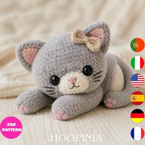 May include: A gray crocheted cat plush toy with a white muzzle, pink inner ears, and pink paw pads. The cat features large black eyes, black embroidered whiskers, and a small beige bow on its head. The text PDF PATTERN and HOOPINIA are visible.
