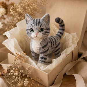 May include: A gray and white crocheted cat figurine with dark gray tabby stripes, green eyes, and a pink nose. The cat is standing upright with its tail curved upward, featuring detailed yarn stitching throughout its body and pointed ears.