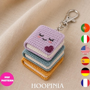 Puede incluir: Un llavero de ganchillo con una pila de tres pequeños libros cuadrados en color lavanda, verde azulado y amarillo mostaza. El libro superior lavanda tiene una cara sonriente bordada y un pequeño corazón morado oscuro. Un cierre de mosquetón de metal plateado.