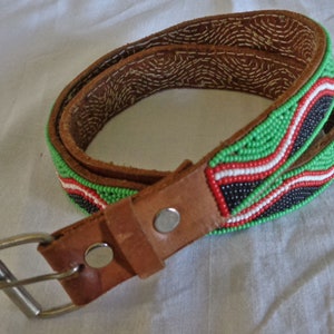 May include: A brown leather belt with a beaded design in green, red, black, and white. The belt has a silver buckle and a leather strap with a stitched pattern. The belt is likely a fashion accessory.
