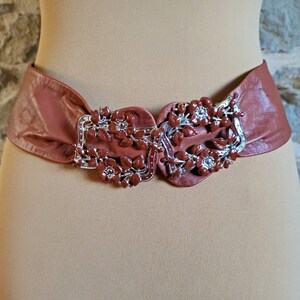 May include: A wide, mauve-colored leather belt with a decorative buckle. The buckle features a silver-toned frame with reddish-brown floral embellishments. The belt is displayed on a mannequin, showcasing its shape and design.