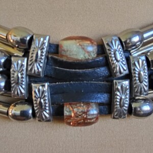 May include: Close-up of a silver-toned necklace with a black leather band. The necklace features decorative silver accents and two rectangular, patterned stones. The necklace has a layered design with multiple silver tubes.