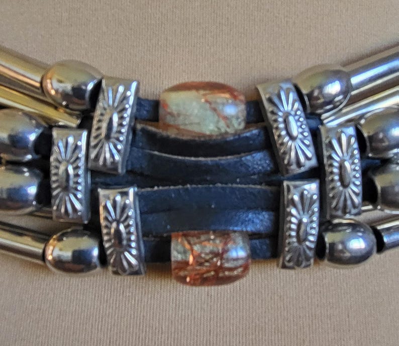 May include: Close-up of a silver-toned necklace with a black leather band. The necklace features decorative silver accents and two rectangular, patterned stones. The necklace has a layered design with multiple silver tubes.