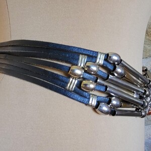May include: A black leather belt with multiple straps and silver-toned metal accents. The belt features cylindrical and rectangular metal details, creating a layered and textured design. The belt is displayed on a mannequin.