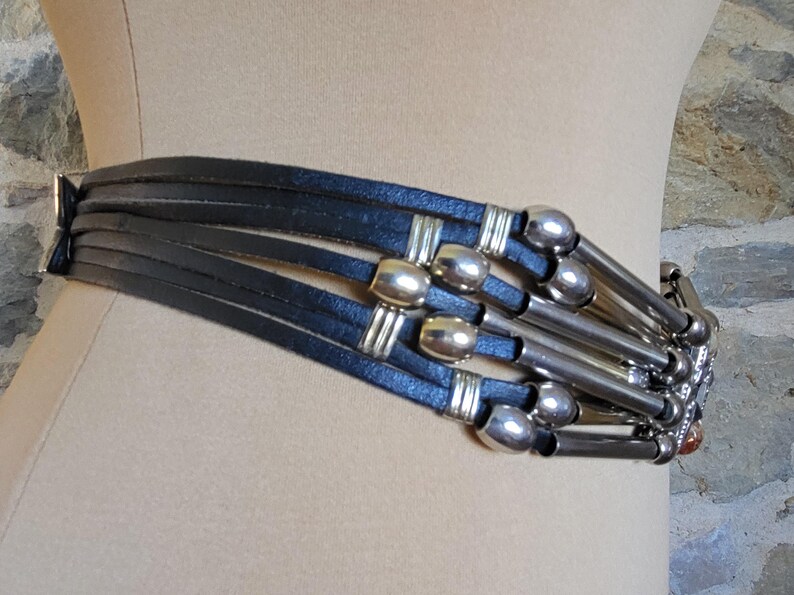 May include: A black leather belt with multiple straps and silver-toned metal accents. The belt features cylindrical and rectangular metal details, creating a layered and textured design. The belt is displayed on a mannequin.