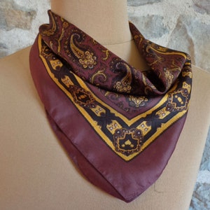 May include: A burgundy paisley patterned scarf with a yellow and black border. The scarf is displayed on a mannequin and has a square shape. The paisley pattern features gold and brown details. The scarf is made of a silky material.
