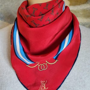 May include: A red silk scarf with a navy blue border. The scarf features a repeating pattern of black equestrian-themed designs, gold rings, and a logo. White and blue stripes are on the side.
