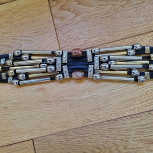 May include: A black belt with silver and gold-toned metal accents. The belt features multiple strands with decorative silver beads and rectangular metal pieces. The center has two decorative stone pieces.