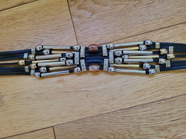 May include: A black belt with silver and gold-toned metal accents. The belt features multiple strands with decorative silver beads and rectangular metal pieces. The center has two decorative stone pieces.