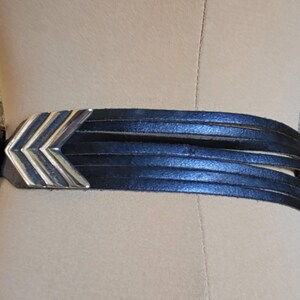 May include: Black leather belt with a silver and gold chevron design buckle. The belt features multiple thin leather straps and a silver buckle closure. The belt is displayed on a mannequin.