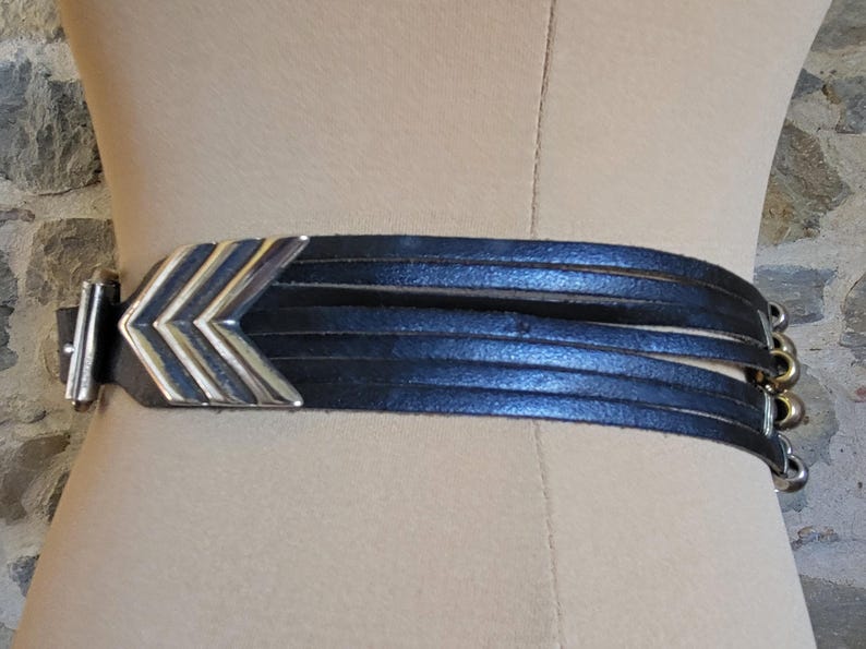 May include: Black leather belt with a silver and gold chevron design buckle. The belt features multiple thin leather straps and a silver buckle closure. The belt is displayed on a mannequin.