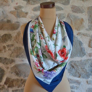 May include: A square silk scarf with a floral print featuring red poppies, purple flowers, and butterflies on a white background. The scarf has a navy blue border and is displayed on a mannequin.