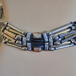 May include: A wide belt featuring silver and gold-toned metal tubes arranged in horizontal rows. The belt has a central rectangular section with decorative metal accents and a dark leather backing. The belt is displayed on a mannequin.