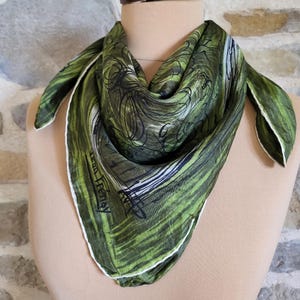 May include: A green silk scarf with a white border and abstract black and white design. The scarf is draped over a mannequin and features the text "Jean Frénay".