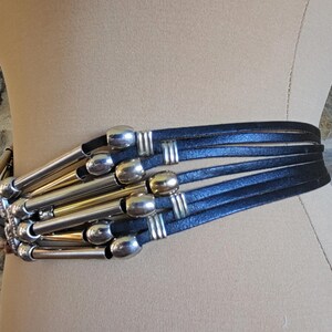 May include: A black leather belt with silver and gold-toned metal accents. The belt features multiple leather straps and decorative metal tubes. The belt is displayed on a mannequin.