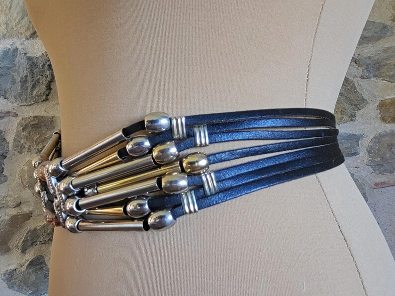 May include: A black leather belt with silver and gold-toned metal accents. The belt features multiple leather straps and decorative metal tubes. The belt is displayed on a mannequin.