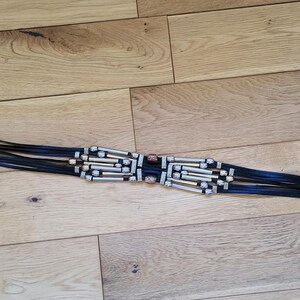 May include: Black belt with silver-tone metal buckle and decorative accents. The belt features multiple black leather straps with silver and gold-tone metal beads and a pointed end with silver chevron details. The belt is approximately 38 inches long.