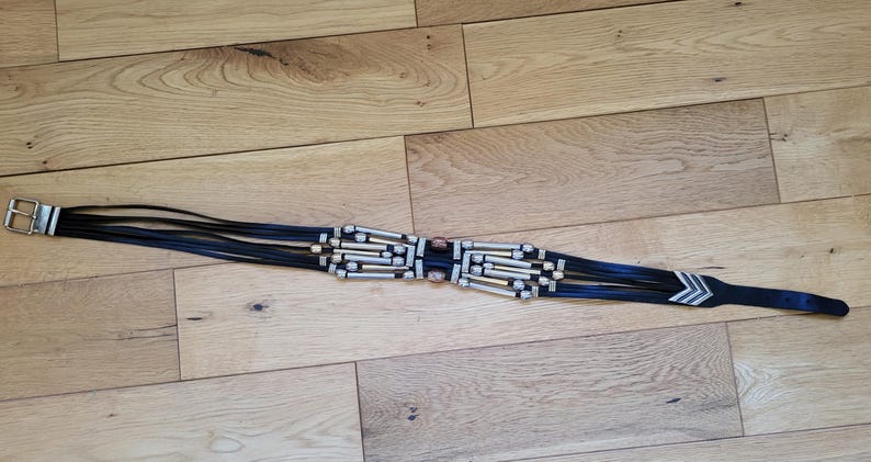 May include: Black belt with silver-tone metal buckle and decorative accents. The belt features multiple black leather straps with silver and gold-tone metal beads and a pointed end with silver chevron details. The belt is approximately 38 inches long.