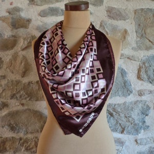 May include: A square silk scarf with a geometric pattern of brown, pink, and white squares. The scarf is displayed on a mannequin and has a dark brown border. The brand name "RODIER" is visible in the corner.