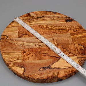 May include: A round olive wood cutting board with a handle. The board is approximately 30 inches in diameter. The board is made of multiple pieces of wood that have been joined together to create a unique pattern.