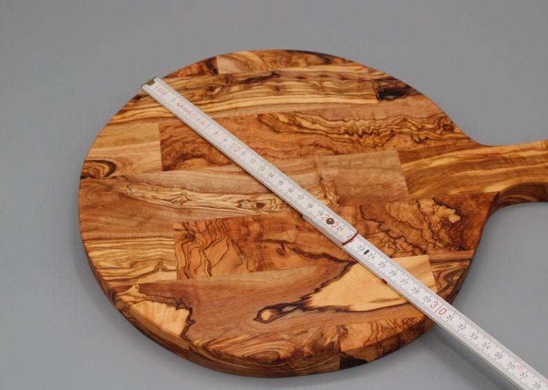 May include: A round olive wood cutting board with a handle. The board is approximately 30 inches in diameter. The board is made of multiple pieces of wood that have been joined together to create a unique pattern.