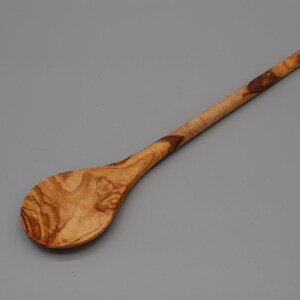 Op de afbeelding: Een houten lepel met een lange steel en een ronde kom. De lepel is gemaakt van olijfhout en heeft een natuurlijke bruine kleur met donkerbruine strepen.