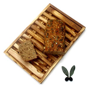 May include: A wooden bread board with a slatted design, featuring a loaf of bread with seeds and a slice of bread on top. The board is made of olive wood and has a natural finish.