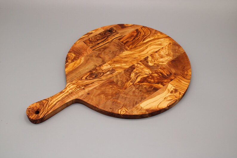 May include: A round olive wood cutting board with a handle. The board has a natural wood grain pattern and is perfect for serving cheese, charcuterie, or other appetizers.