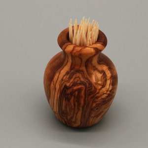 May include: A small, wooden, olive wood toothpick holder in the shape of a vase. The holder is filled with toothpicks.