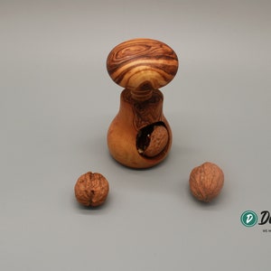 Peut inclure: Un casse-noisette en bois en forme de champignon avec un motif de grain de bois brun et blanc. Trois noix sont sur une surface grise, l'une est partiellement cassée dans le casse-noisette.
