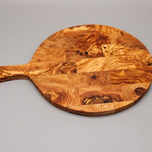 May include: A round olive wood cutting board with a handle. The board is made of multiple pieces of wood that have been joined together to create a unique pattern.
