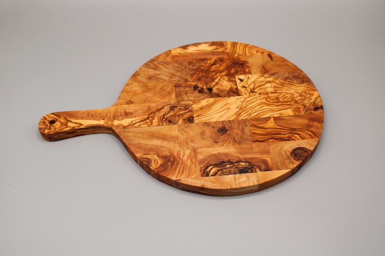 May include: A round olive wood cutting board with a handle. The board is made of multiple pieces of wood that have been joined together to create a unique pattern.