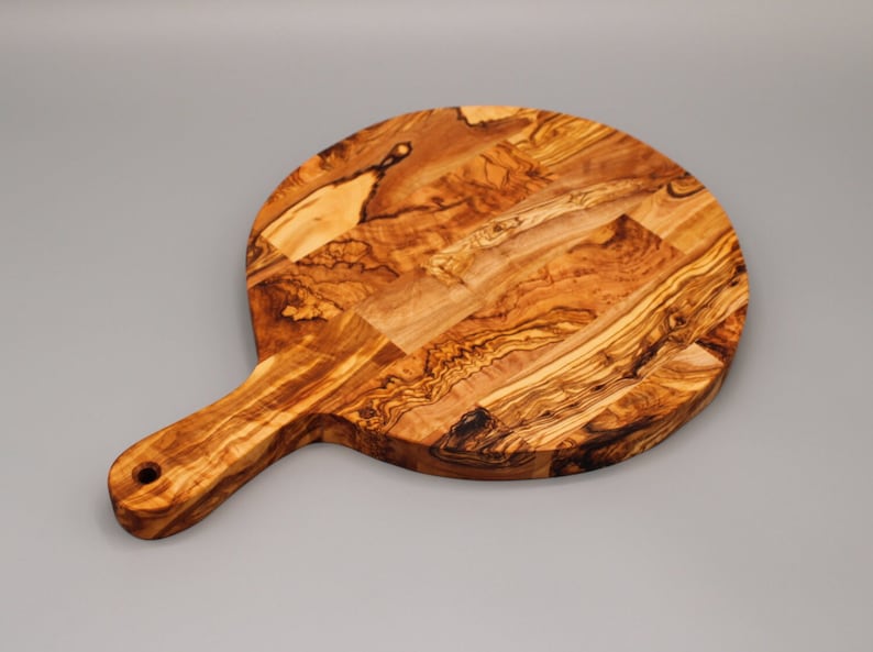 May include: A round olive wood cutting board with a handle. The board has a natural wood grain pattern and is perfect for serving cheese, charcuterie, or other appetizers.