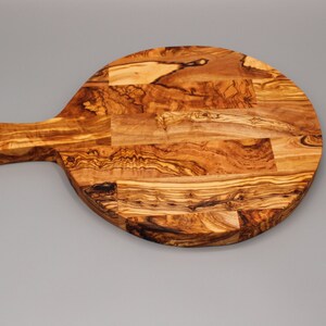May include: A round olive wood cutting board with a long handle. The board is made of multiple pieces of wood that have been joined together to create a unique pattern.
