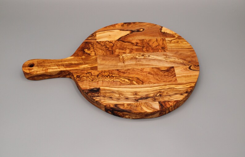 May include: A round olive wood cutting board with a long handle. The board is made of multiple pieces of wood that have been joined together to create a unique pattern.
