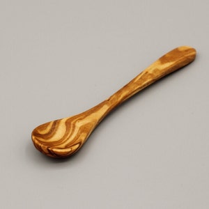 May include: A small, wooden spoon with a natural wood grain pattern. The spoon is made of olive wood and has a rounded bowl.