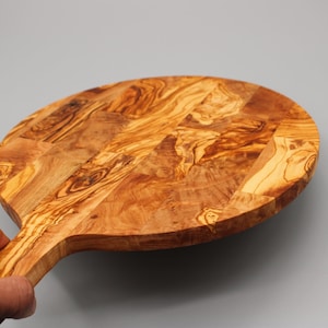 May include: A round olive wood cutting board with a handle. The board has a natural wood grain pattern and is made of a single piece of wood.