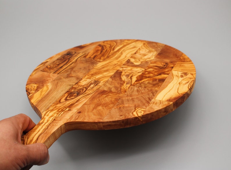 May include: A round olive wood cutting board with a handle. The board has a natural wood grain pattern and is made of a single piece of wood.
