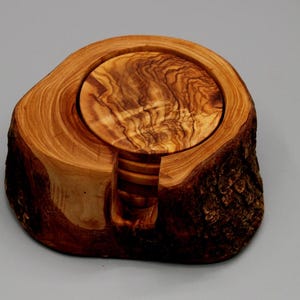 Può includere: Un set di quattro sottobicchieri in legno di ulivo in un supporto in legno naturale. I sottobicchieri sono rotondi e hanno una finitura rustica e naturale. Il supporto è realizzato in un pezzo di legno di ulivo con la corteccia ancora attaccata.