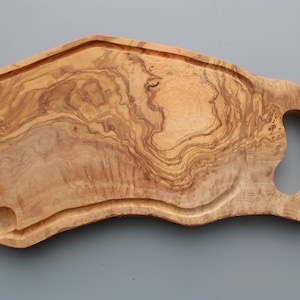 Puede incluir: Una tabla de servir de madera natural grande con una forma única y libre y un asa. La tabla tiene un rico color marrón con un hermoso patrón de veta.