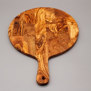 May include: A round olive wood cutting board with a handle. The board is made of multiple pieces of wood that have been joined together to create a unique pattern.